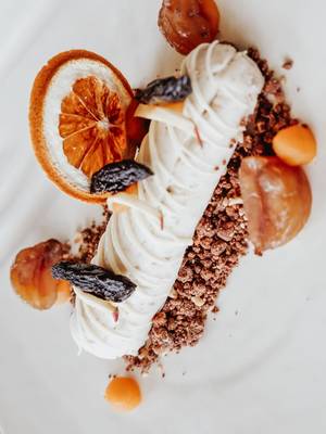 Close-up of a plated, gourmet dessert on a white plate, featuring a white cream roll over chocolate crumbles, garnished with a dried orange slice and candied chestnuts.